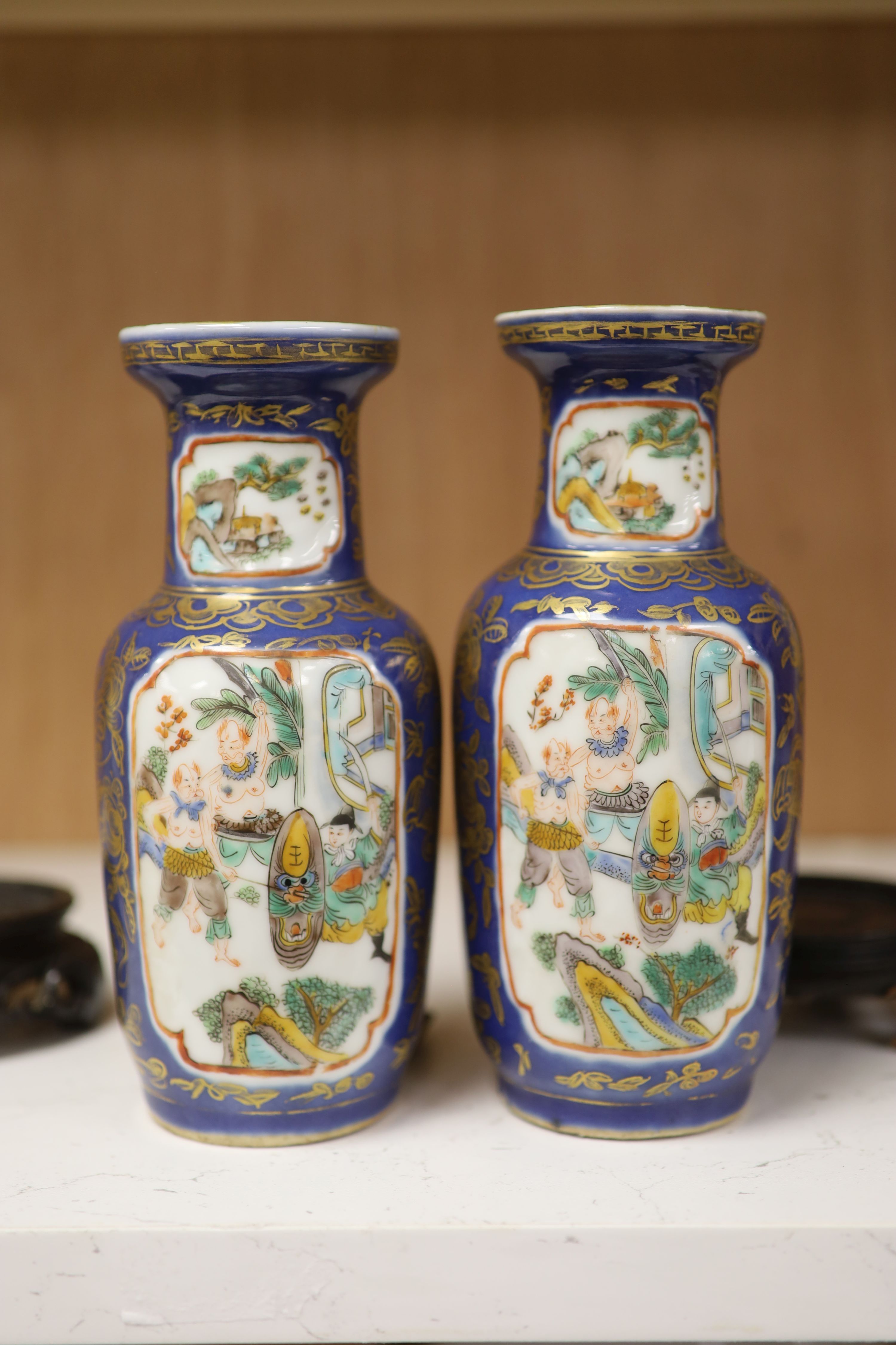 A pair of Chinese Kangxi style blue-ground vases, late 19th century decorated with panels of warriors and heightened in gilt, on carved hardwood stands, height 18cm excluding stand